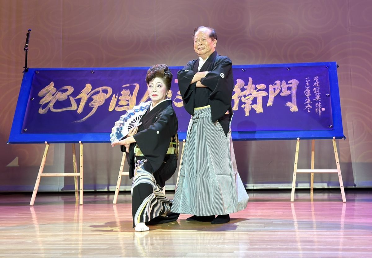 圖／新邦樂舞踊若泉流台灣分家家元若泉德玲（林汶玲老師，前）於舞台上優雅展演日本傳統舞踊之美，展現深厚藝境與文化風韻。後方為林暉博士親題贈書〈紀伊國若泉門〉，象徵日台藝術薪傳與文化交流的深厚情誼。（必紅演藝 提供）